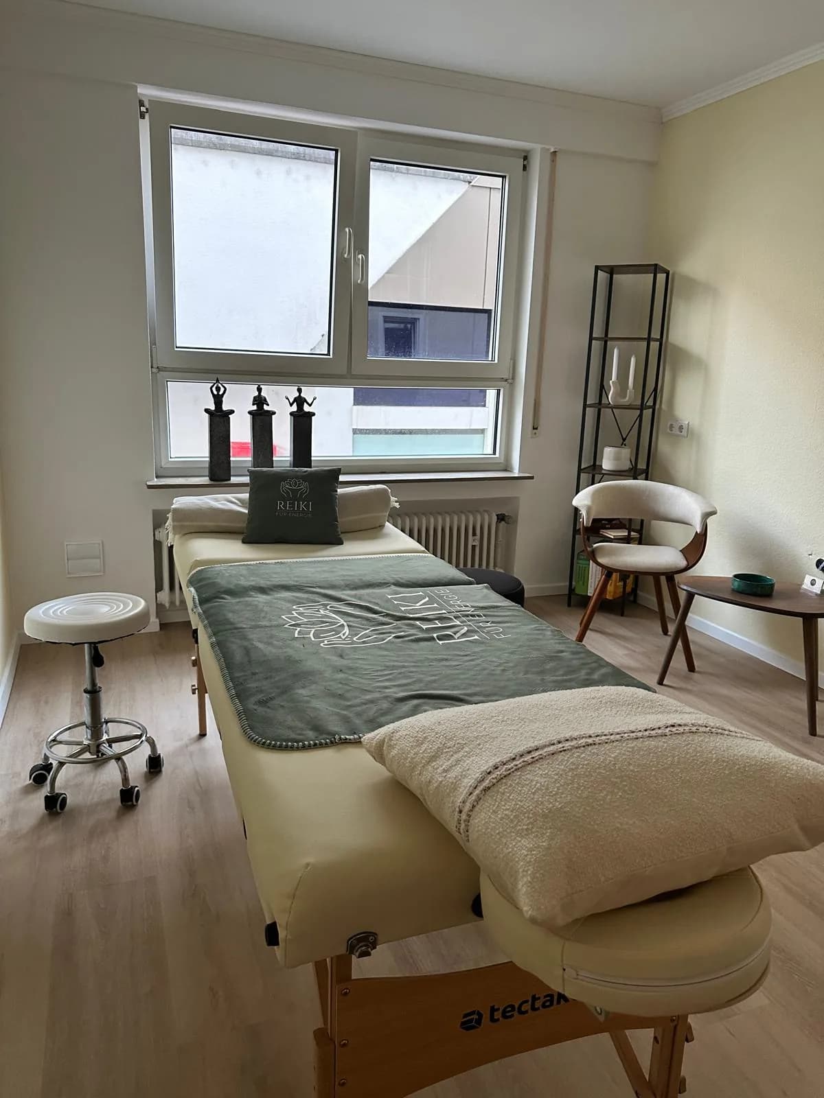 Der Praxisraum: Liege mit Salbeidecke und Reiki-Kissen, Fenster, Sessel und Regal